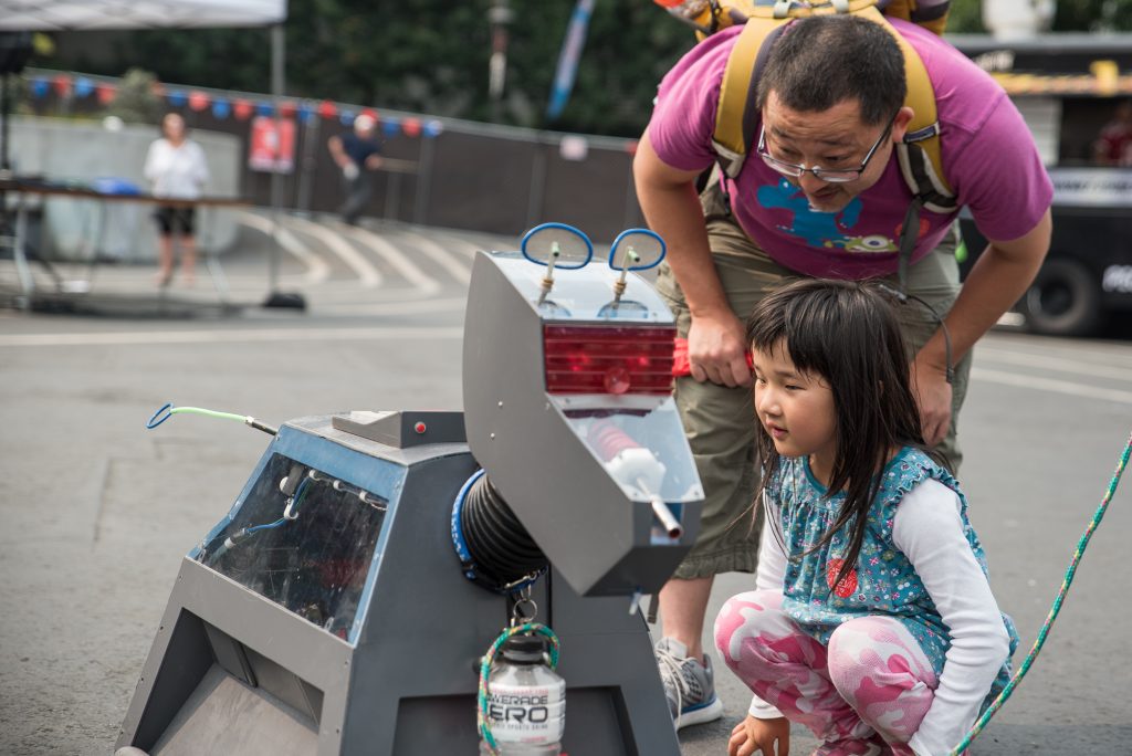 A young girl and her father examine a robot dog art piece. 