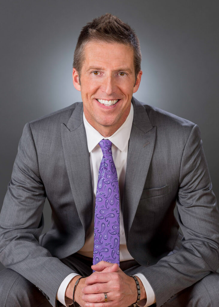 Photo of a white man in a business suit with a purple tie sitting down and smiling