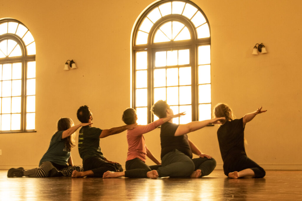 A group of dancers sit in a light filled room with their backs to the camera.