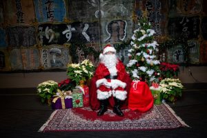 An Asian American person, dressed as Santa Claus, sits in front of a decorated Christmas tree surrounded by colorfully wrapped presents.