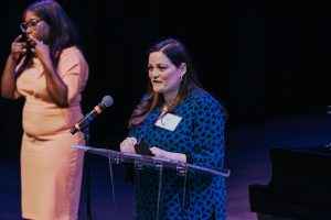 A woman wearing a blue shirt speaking at a podium with an ASL interpreter standing in the background.