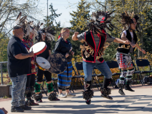 A group of people dressed in traditional indigenous garments standing outside and in front of a line of chairs. Two people are jumping, and four others are seen singing and playing instruments.