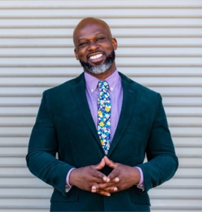 Marc is a Black man with bald head and a salt and pepper beard. He is smiling in a suit and tie, looking at the camera in front of a gray background.