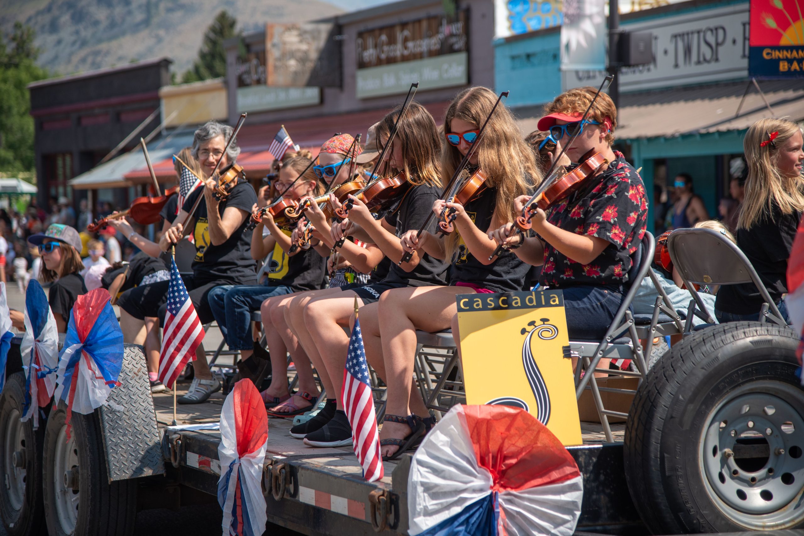 In the middle of a parade, a line of children sitting in chairs with sunglasses play the violin for the crowd.