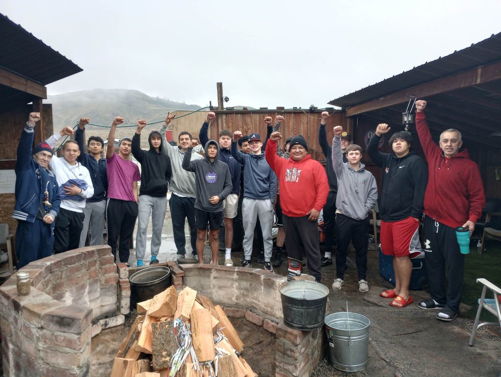 Tucked away in a cloudy landscape are a group of people standing outside and in front of a fire pit. Everyone is facing the camera and holding a fist in the air.