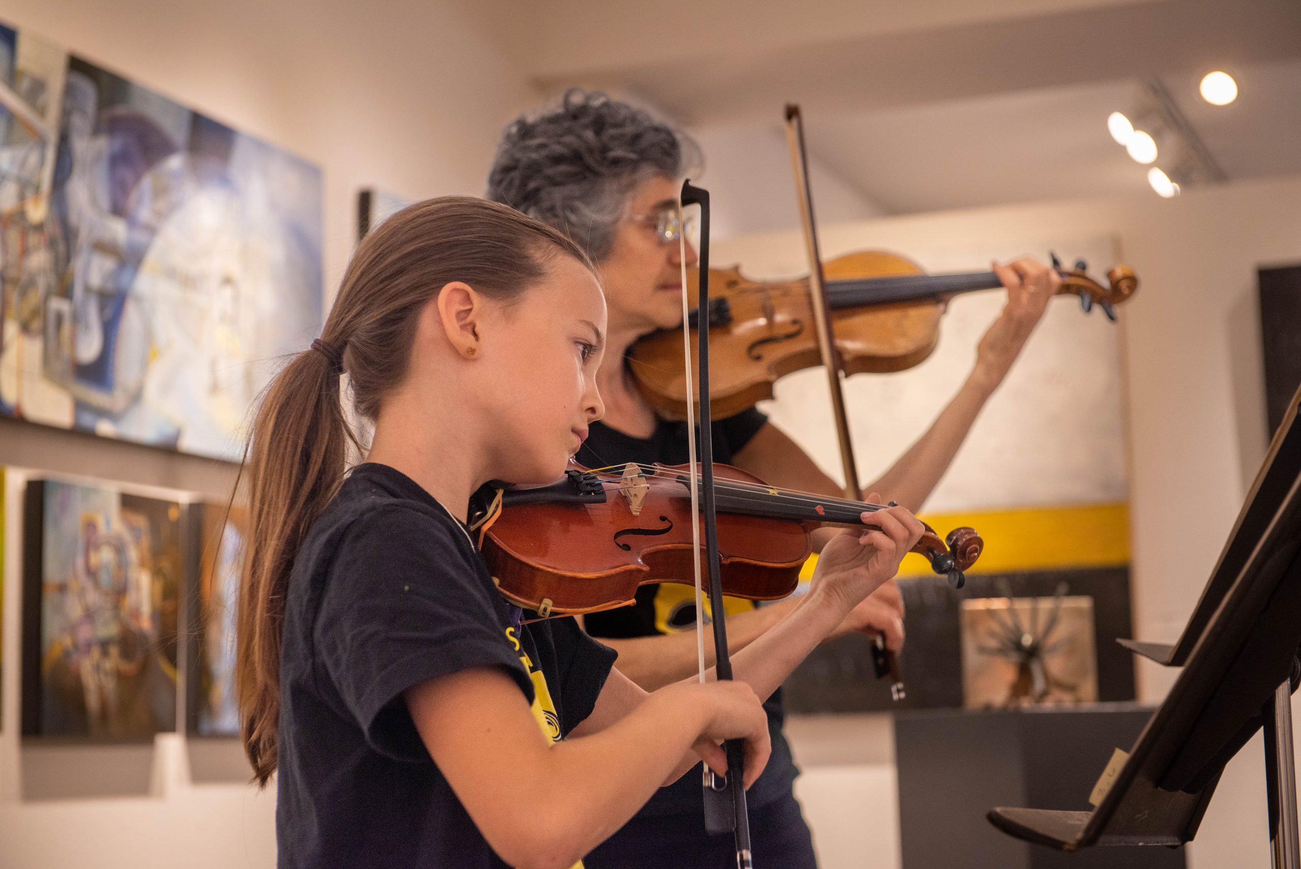 In an art gallery, two people are playing the violin. The younger one is in the foreground with long brown hair and a black T-shirt, looking at a sheet of music. The older person has salt and pepper short hair and glasses and is also wearing black.