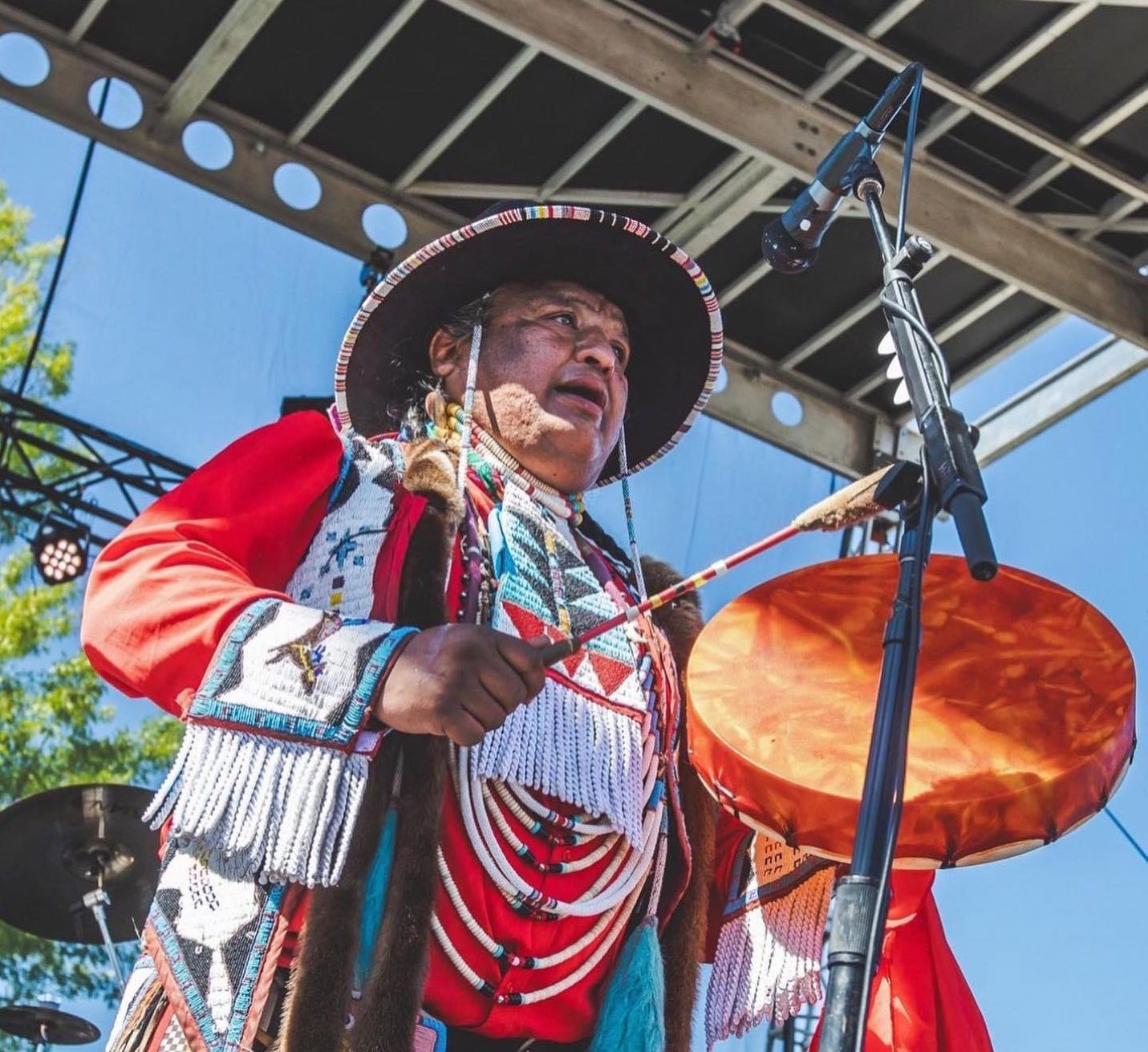 Pictured is Dan Nanamkin performing during a sunny day. He is wearing traditional Indigenous clothing, and playing an indigenous instrument as he sings into the microphone.