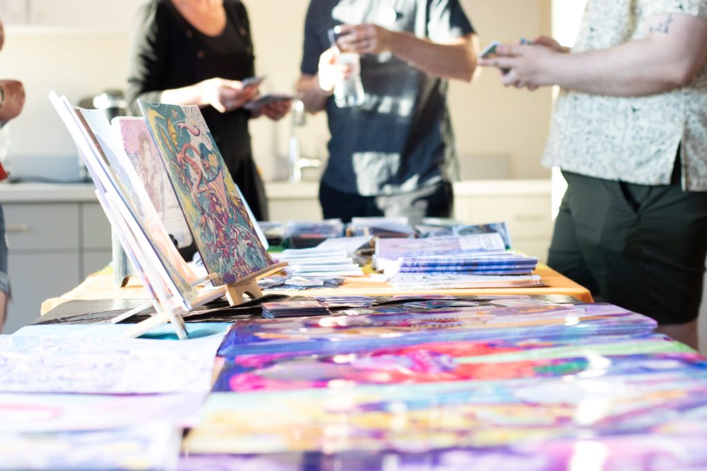 Looking down at the long edge of a table filled with booklets and pamphlets. Two people are the furthest in the background and a third person standing in front of the table in the foreground.