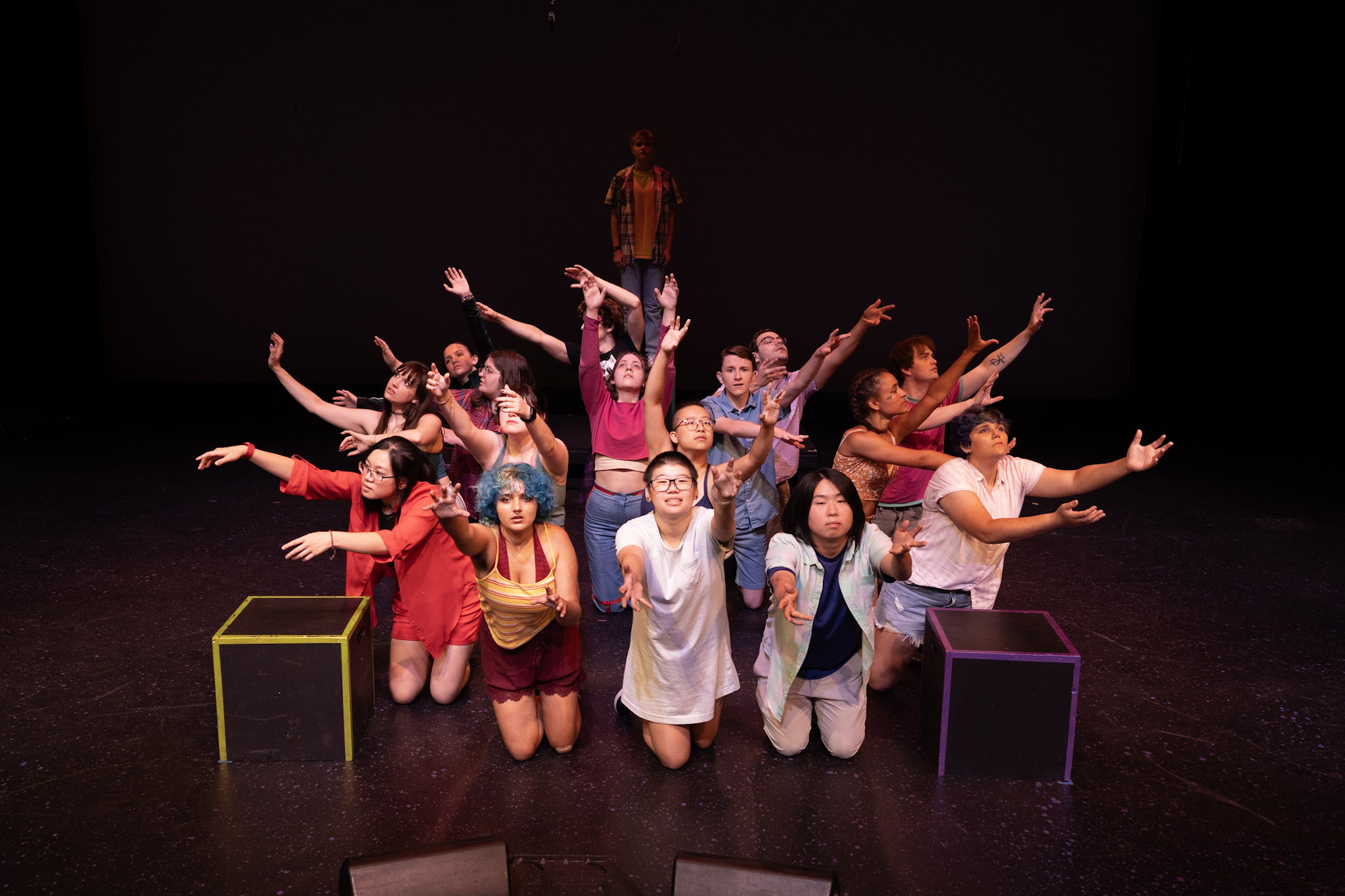 Around 10-15 performers arranged in a central location on a theater stage spoke outwards with their arms stretch outward and away from the center.