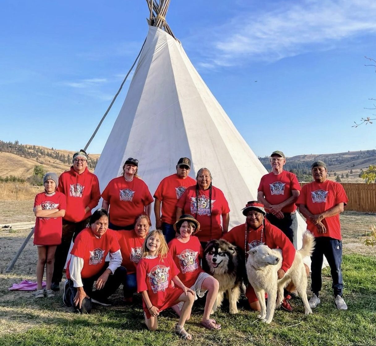 on a sunny day with blue skies and very few clouds a group of folks in a retd t-shirt all smile towards the camera for a group photo. Behind them is an indigenous structure.