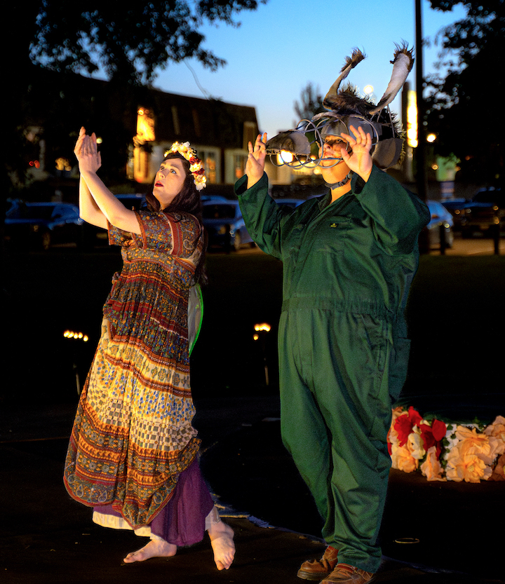 Two actors are standing outside dressed in their costumes as the sun goes down. They are both gesturing with their arms raised towards their left side.