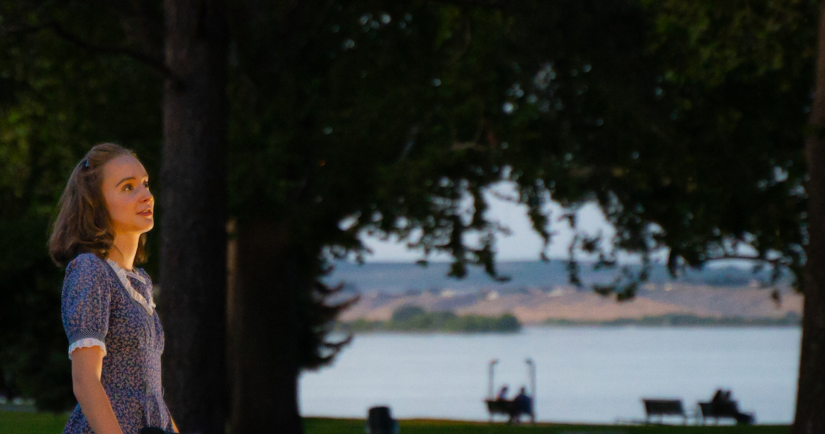 The image is set outside in the forest overlooking a lake. You can see park benches and two people in the background. One actress is wearing a blue floral dress looking in the upper right corner of the screen with a slight smile.