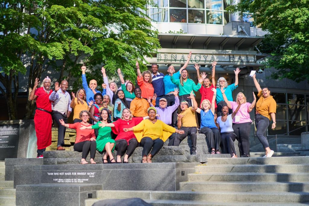 A group of about 20 people in front of Benaroya Hall. Each person is wearing a shirt that is a bold gold, teal, red, green or blue with black pants. Everyone is smiling with their arms reached out above them in joy.