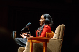 A woman in sitting on stage in a tan arm chair in front of a microphone.