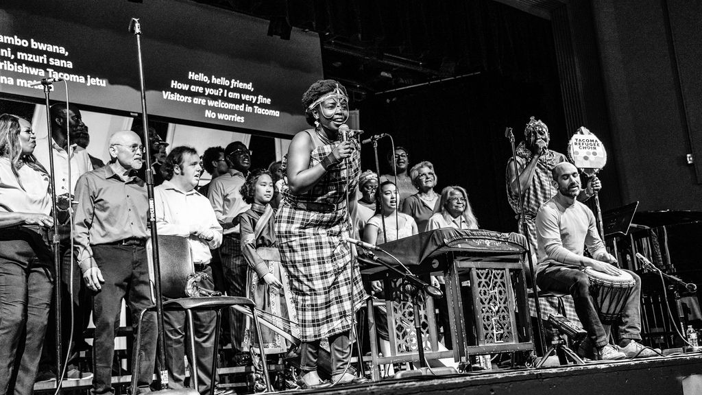 A black an white image of a diverse group of people on state. Some are singing and some are playing instruments.