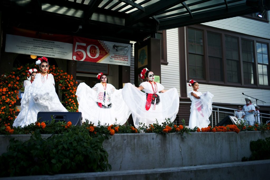 A group of dancers in long white and red costumes and makeup, dancing and performing on stage.