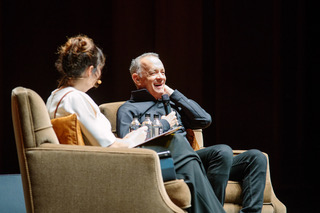 A man and woman sit in tan armchairs with mics on a stage in mid conversation.