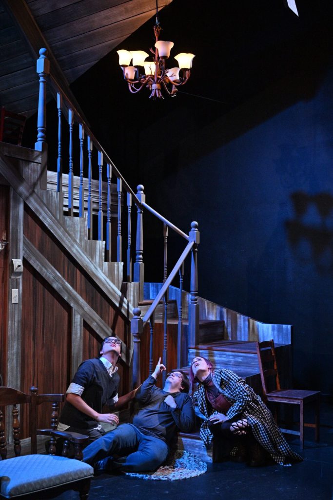 Three actors on a the set of a house. All three are at the bottom of a staircase lying down and looking up towards the ceiling.