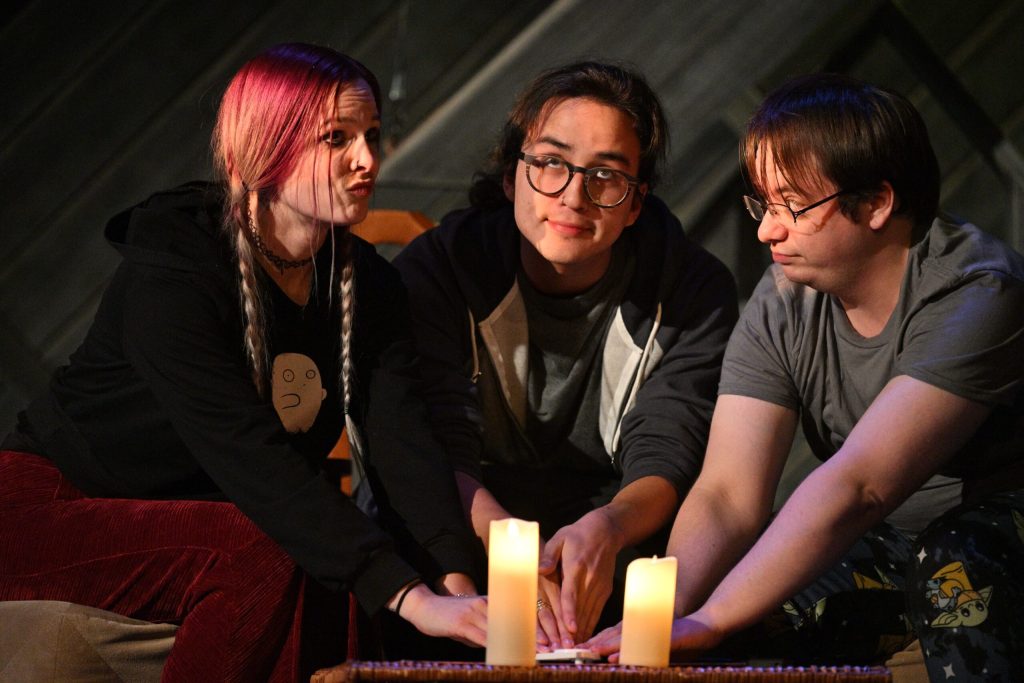 A group of actors on stage sitting in chair and looking in different directions with their hands on what could be a ouija board.