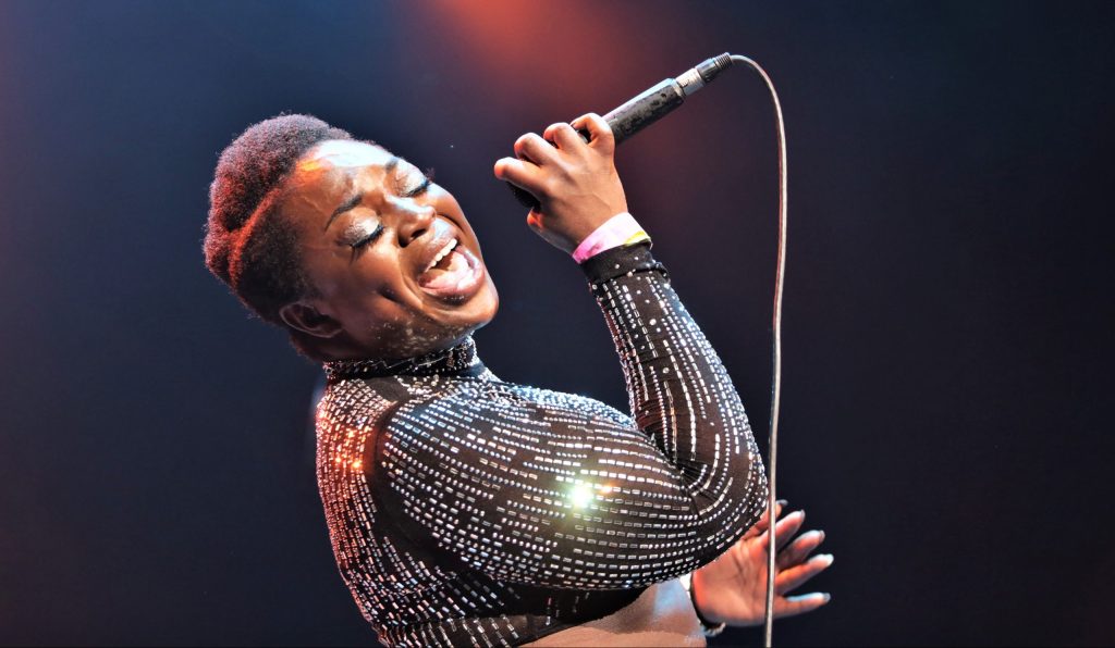 Shaina Shepard is wearing a black long sleeve sequined top with short black hair, leaning back and singing into the microphone.