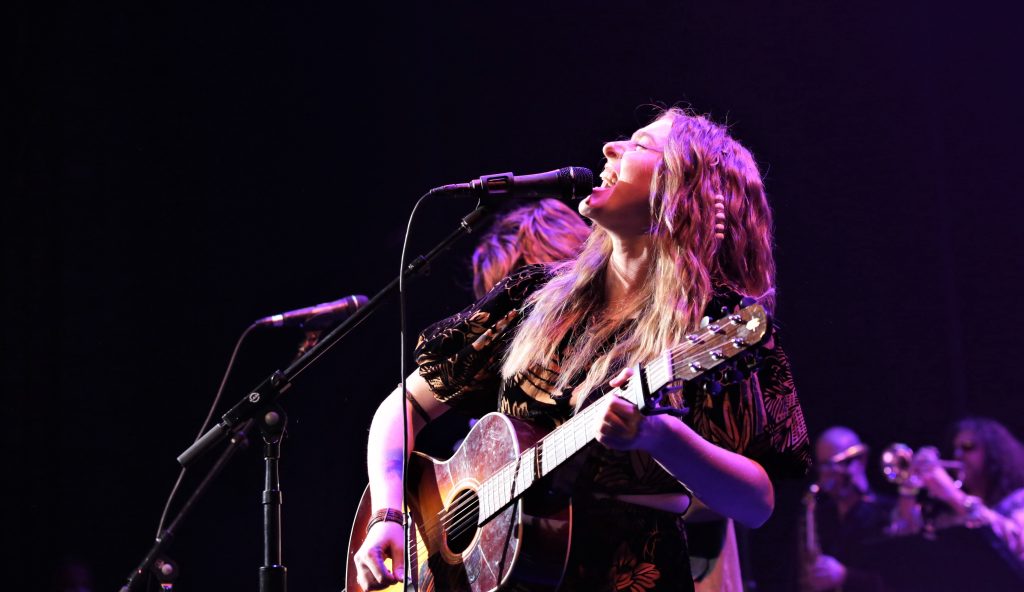 The performer in this image is holding a guitar, has long blond hair and is singing into the microphone looking up to the sky.