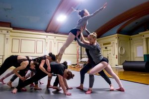 A group of dancers in a studio. The majority of the dancers are supporting one of the dancers in a lift so lifted dancer can reach their arm out towards the sky.