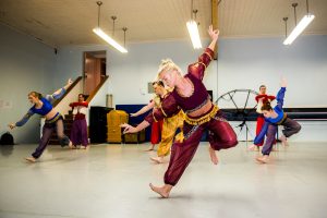 A group of dancers in a studio mid-movement in red, purple and yellow costumes.