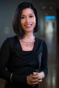 Mari is standing in front of the camera smiling with hands clasped in front of them. Mari has short dark brown, shoulder-length hair, a silver necklace and a long sleeve black shirt.