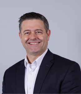 Bill Chapin has short black hair and is wearing a black jacket and a white button-up shirt. He is also smiling at the camera.