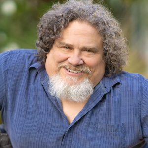 Jim has short curly grey hair and a beard. He is wearing a blue button-up shirt and is smiling at the camera.