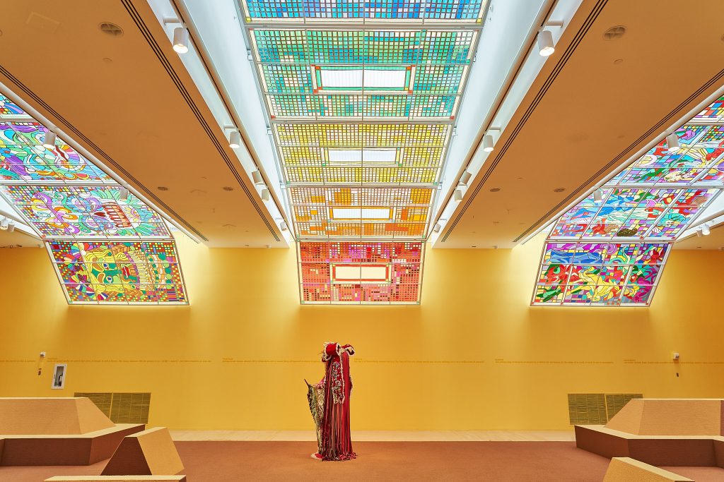 An Indigenous man wearing tribal clothing stands in the middle of a room that's decorated with beautiful tiles on the ceiling. The tiles are rainbow-colored and glass-stained.