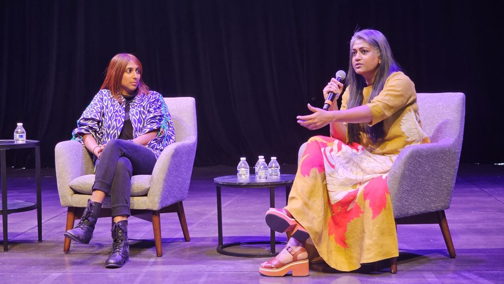 Two Southeast Asian women from Tasveer are giving a talk on stage. One of them is wearing a blue blazer and jeans. The other women is wearing a burnt orange dress with red and cream colored flowers on it.