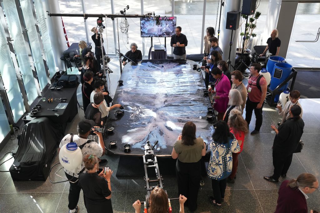 A crowd of attendees gather around a live painting with artist Jesse Higman. The table has splashes of blue, purple, and white on it.