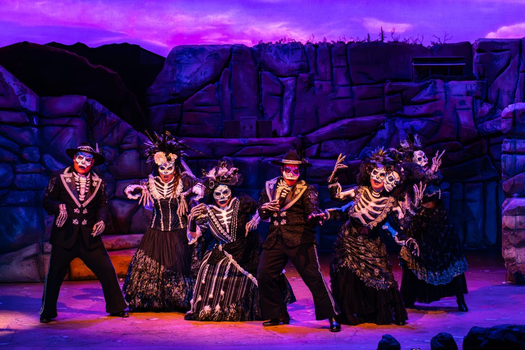 Seven performers with paint on their faces resembling skulls, using vibrant colors, intricate designs, and floral decorations perform on a stage.