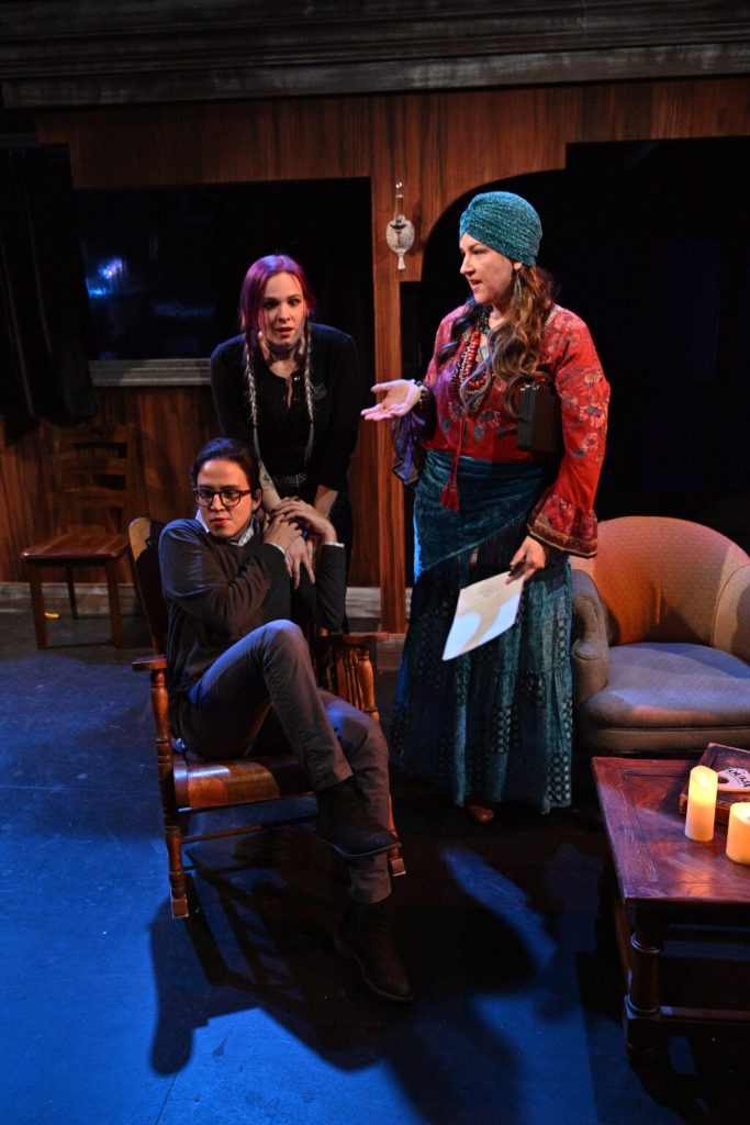 Three actors from the Sound Theatre are performing in a play. One woman in pigtails is holding the hands to a woman in distress who's sitting in a chair. The third woman is standing and wearing clothes similar to a fortune teller.