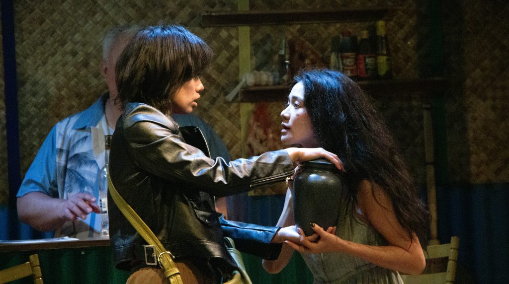 Two Asian actors gaze into each other's eyes on a stage. One is wearing a leather jacket and the other holds the helmet the other person is carrying.