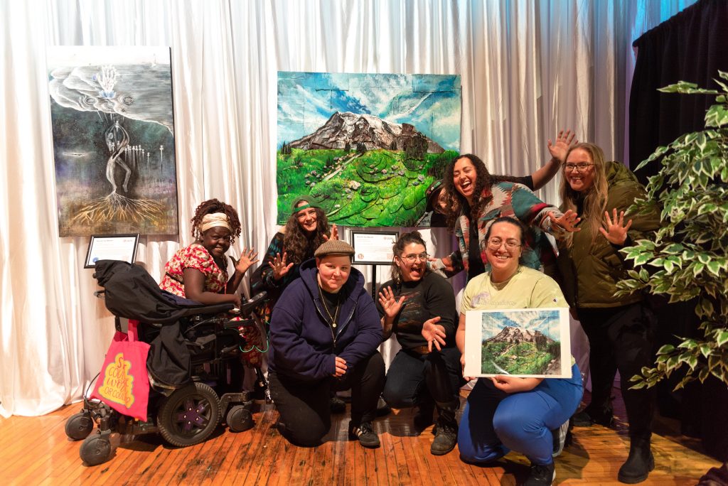 Seven artists pose in front of a painting of a mountain.