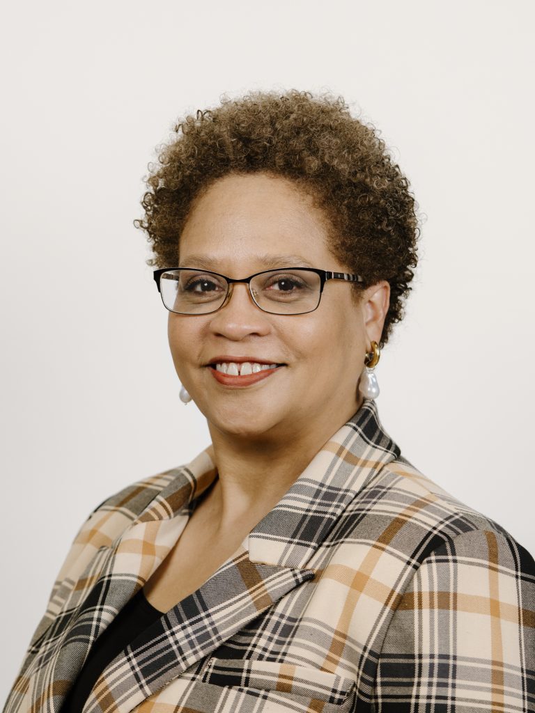 Maria Kolbe-Wolfe is a Black woman with curly brown hair, thin-rimmed glasses wears a plaid sweater over a black undershirt. She's also wearing pearl earrings.