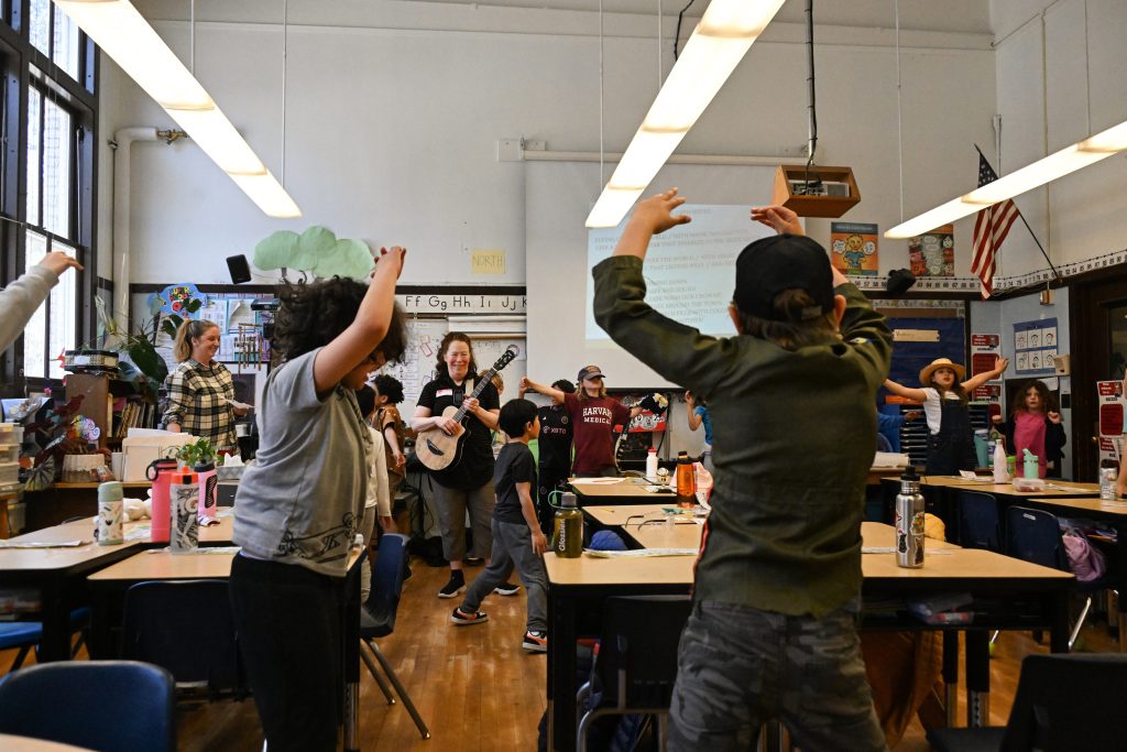 A classroom full of youth raise their hands in the air as someone plays the guitar.