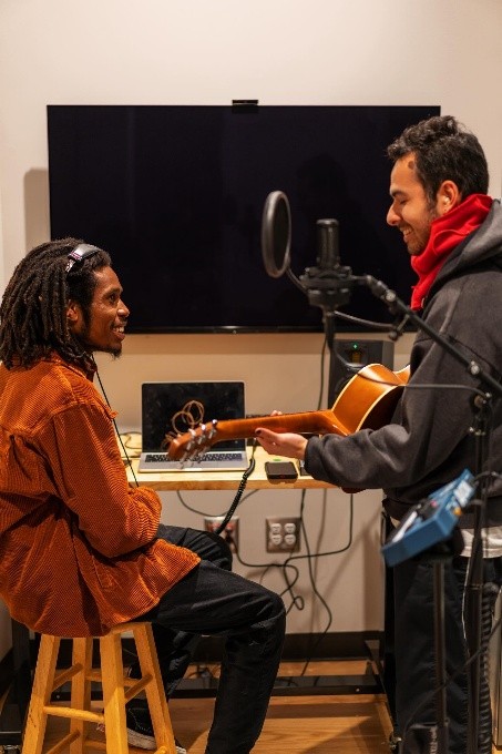 A black man wearing headphones, an orange sweater and black pants sits on a stool in a recording studio with another young man who's playing a guitar. The room is filled with a large mounted television screen, laptop, and other recording equipment.
