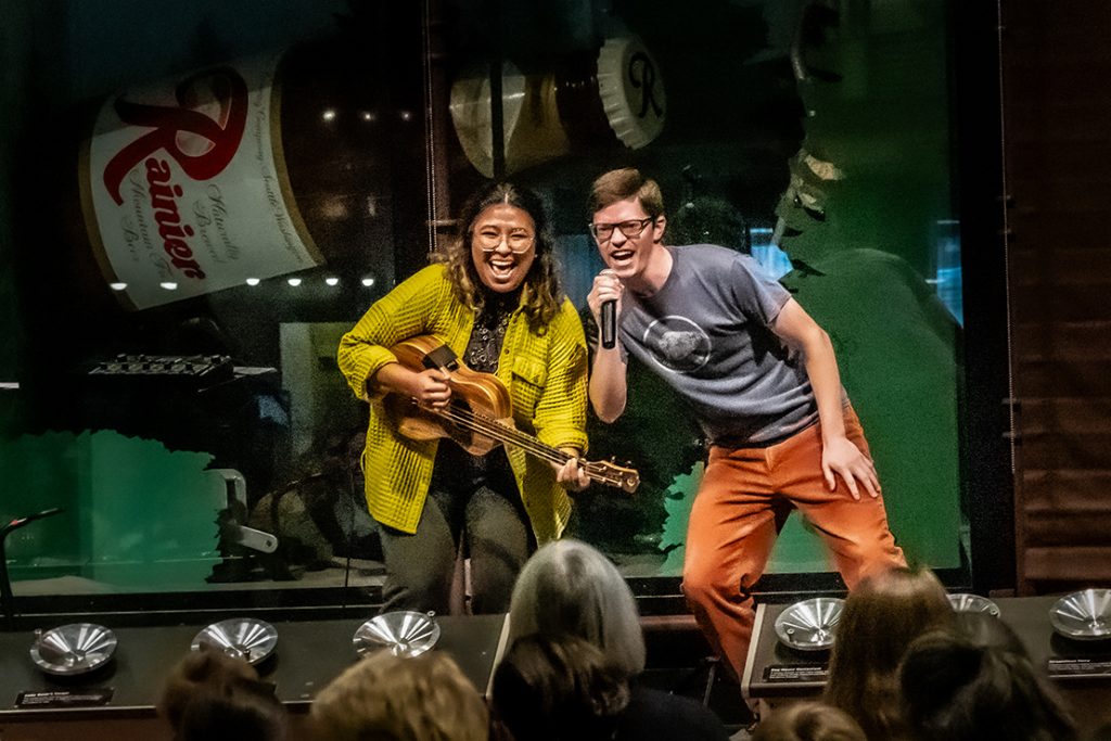 A woman plays a guitar, singing, alongside a man.