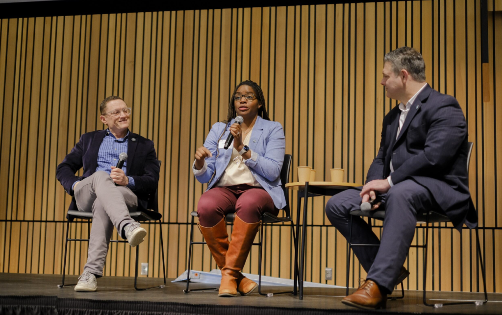 A panel of professionals, two white men and a Black woman, speak into a mic with a brown backdrop.