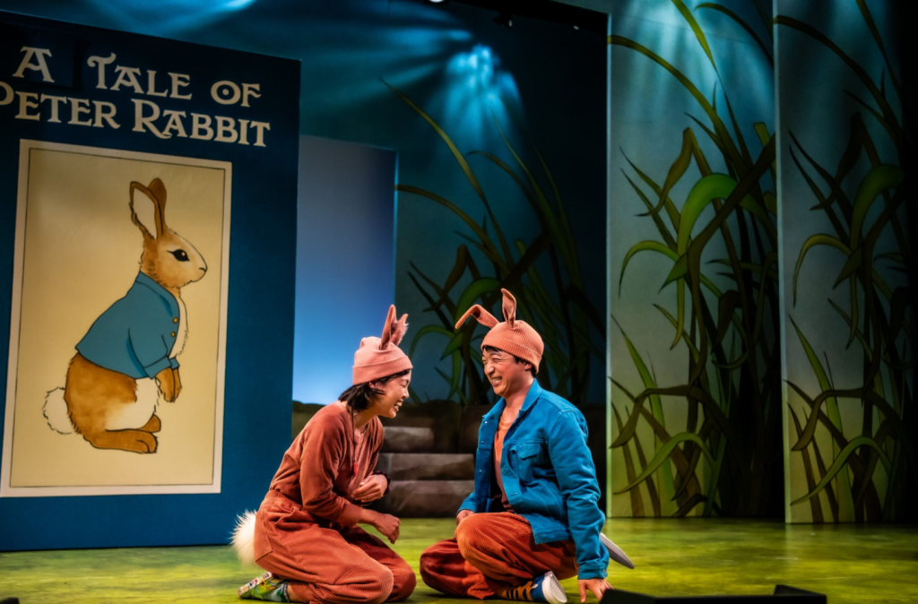 A woman and a man are performing The Tale of Peter Rabbit. They are dressed as bunnies and the man is also wearing Peter Rabbit's signature blue cardigan. A giant book of the Tale of Peter Rabbit and structures with grass-like pictures are in the background.