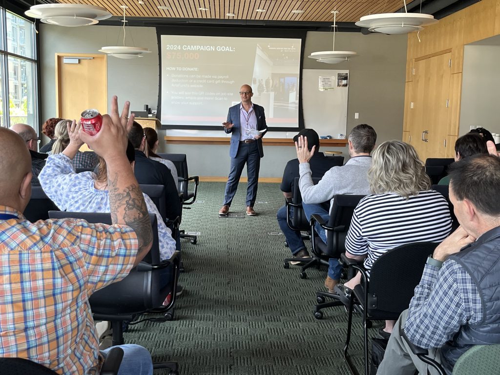 An adult man wearing a blue blazer over a light-blue shirt and blue slacks gives a presentation in a conference room in front of a group of professionals. Behind him is a screen with the words, "2024 Campaign Goal: $75,000"