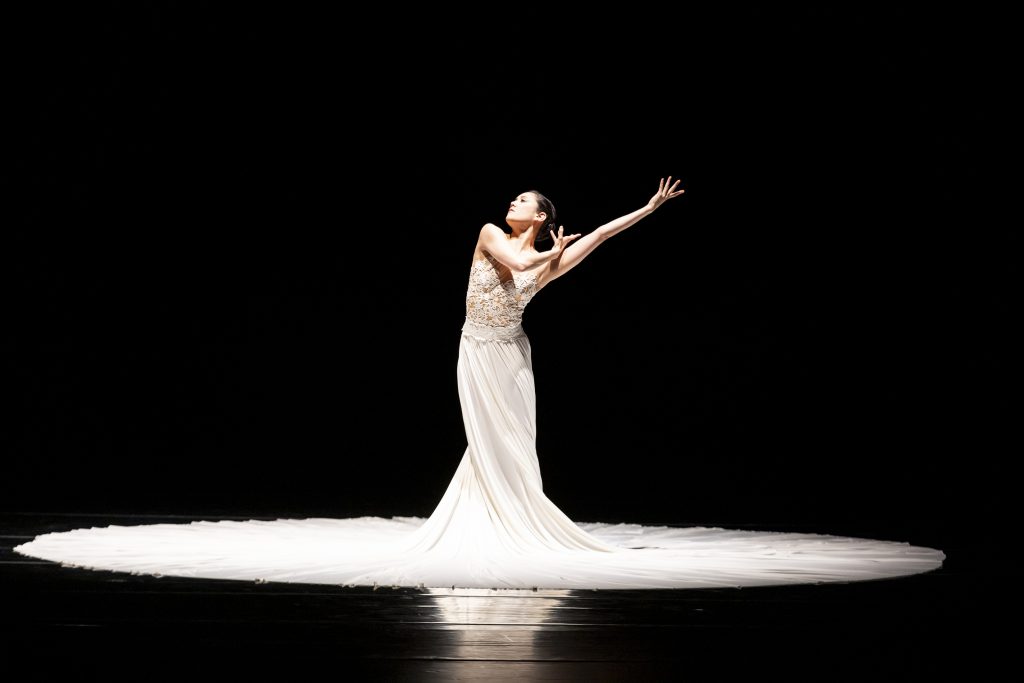 A ballet performer poses elegantly in a white gown against a black background on stage.