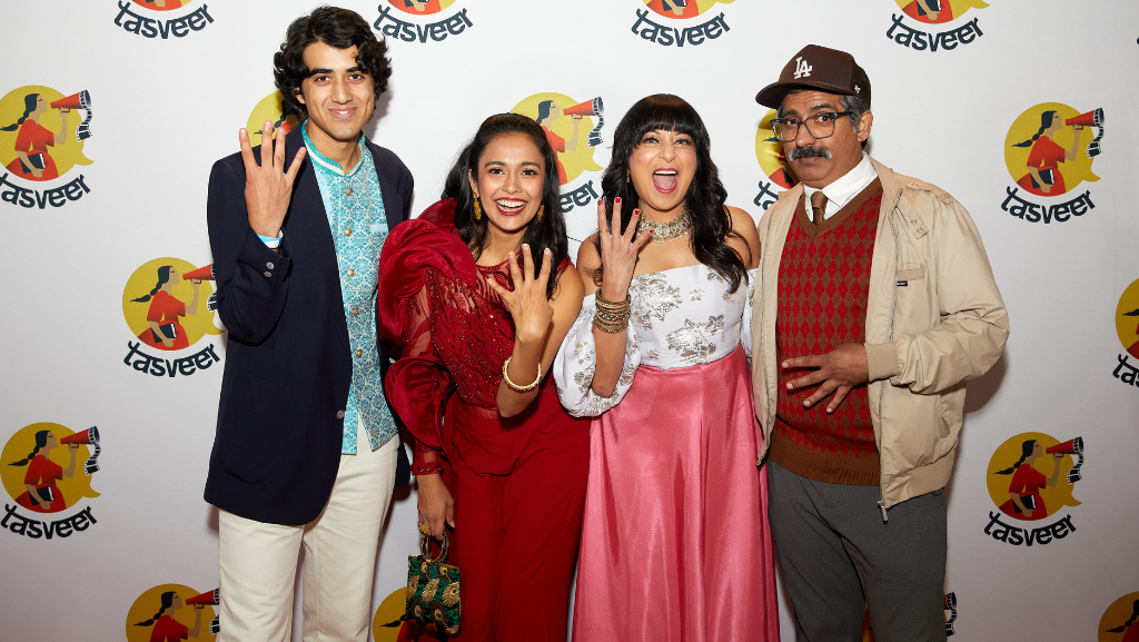 Four TASVEER filmmakers stand in front of a backgdrop with the Tasveer logo spread across it. They are all Southeast Asian, dressed nice, and holding up four fingers.