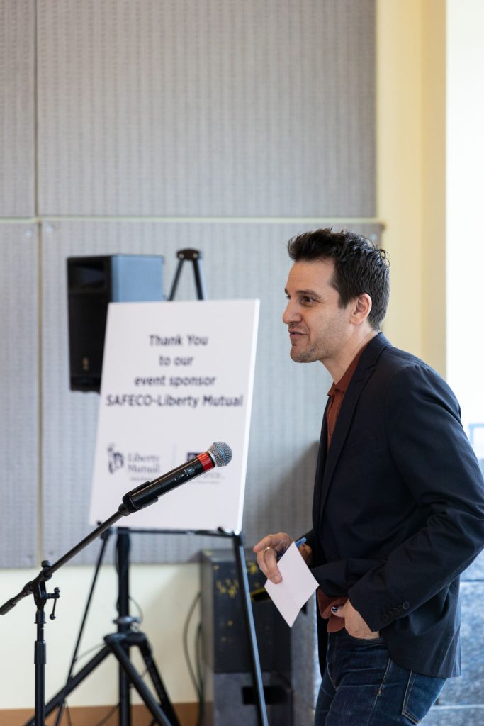 An adult man stands in front of a mic. He's wearing a navy-clue cardigan over an orange button-up shirt, and blue jeans. Behind is is a sign reading, "Thank You to our event sponsor SAFECO - Liberty Mutual"