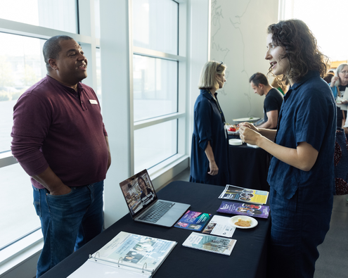 At an ArtsFund Board Matching event, a young professional talks to a presenter, inquiring about their organization. The table in front of them has a laptop and materials from their organization.