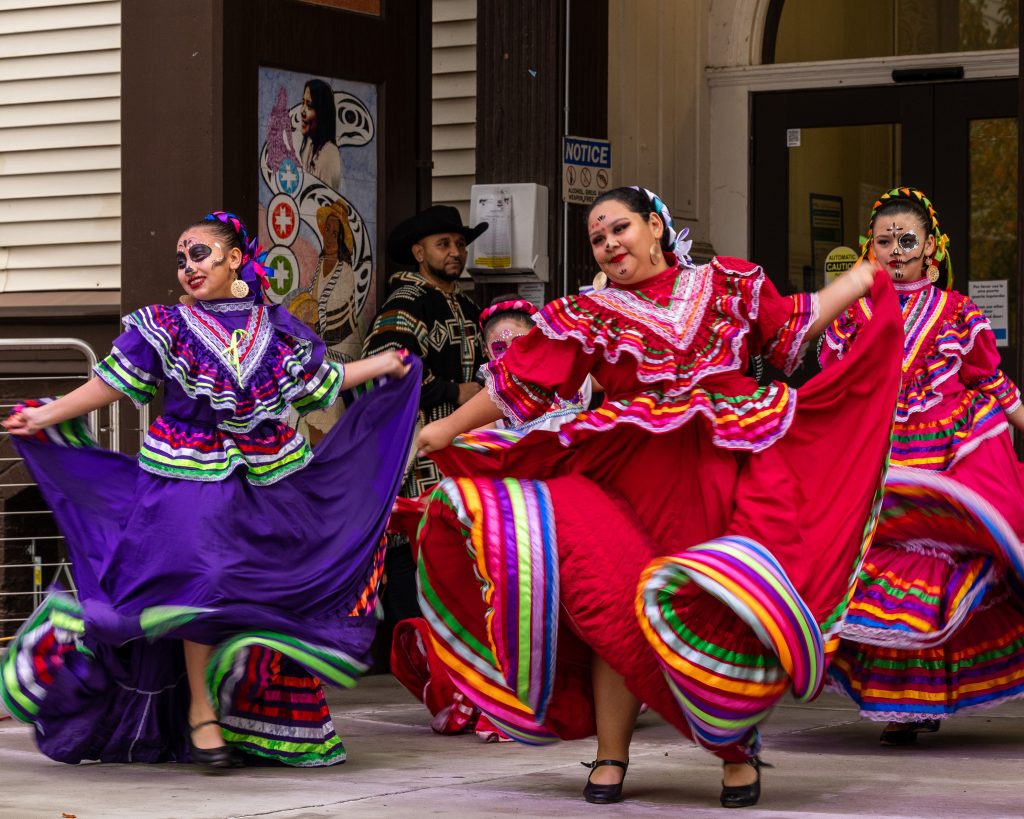 Beautiful performers dance at El Centro de la Raza. They are dressed in bright red and purple, all of whom are wearing makeup resembling skulls.