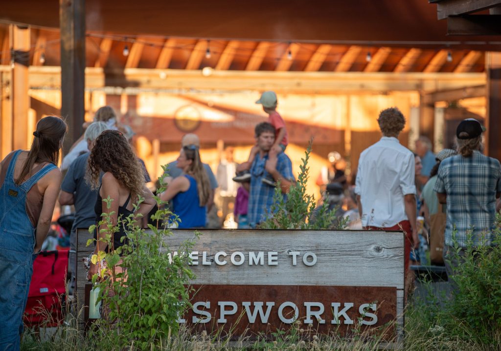 Crowd of people socializing outdoors near a wooden sign that reads 'Welcome to TrispWorks' under a covered area.